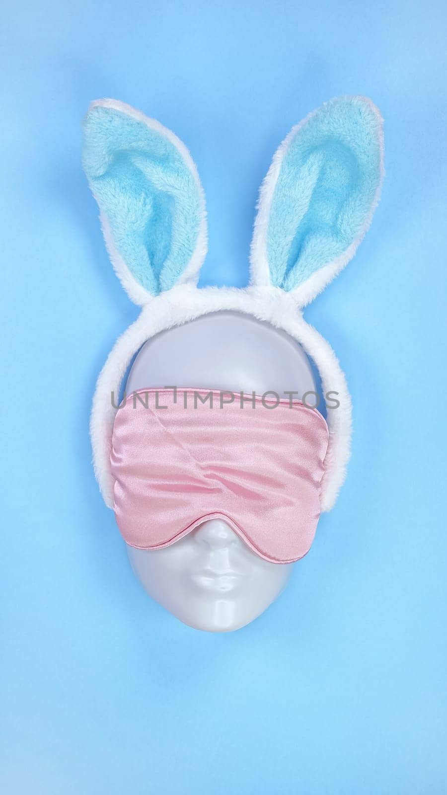 Pink sleeping eye mask on mannequin face with blue fluffy Easter bunny ears on blue background, sleeping disorder. Holidays, Head accessory. Plastic face. by JuliaDorian