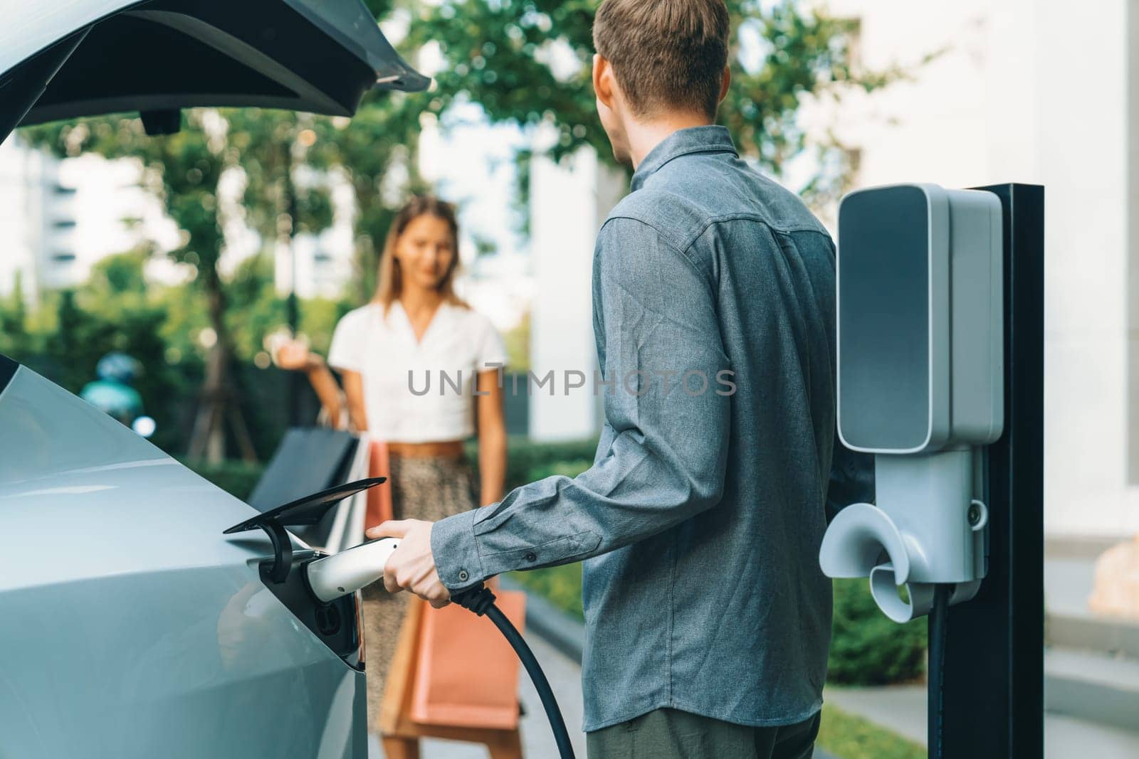 Young couple travel with EV electric car charging in green sustainable city outdoor garden in summer shows urban sustainability lifestyle by green clean rechargeable energy of electric vehicle innards