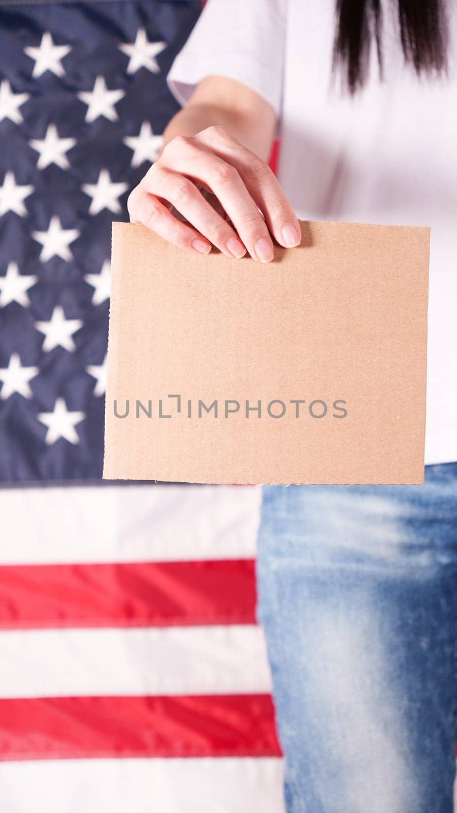 Young woman holds empty cardboard with Space for Text sign against American flag on background. Girl protesting anti-abortion laws. Feminist power. Womens rights freedom