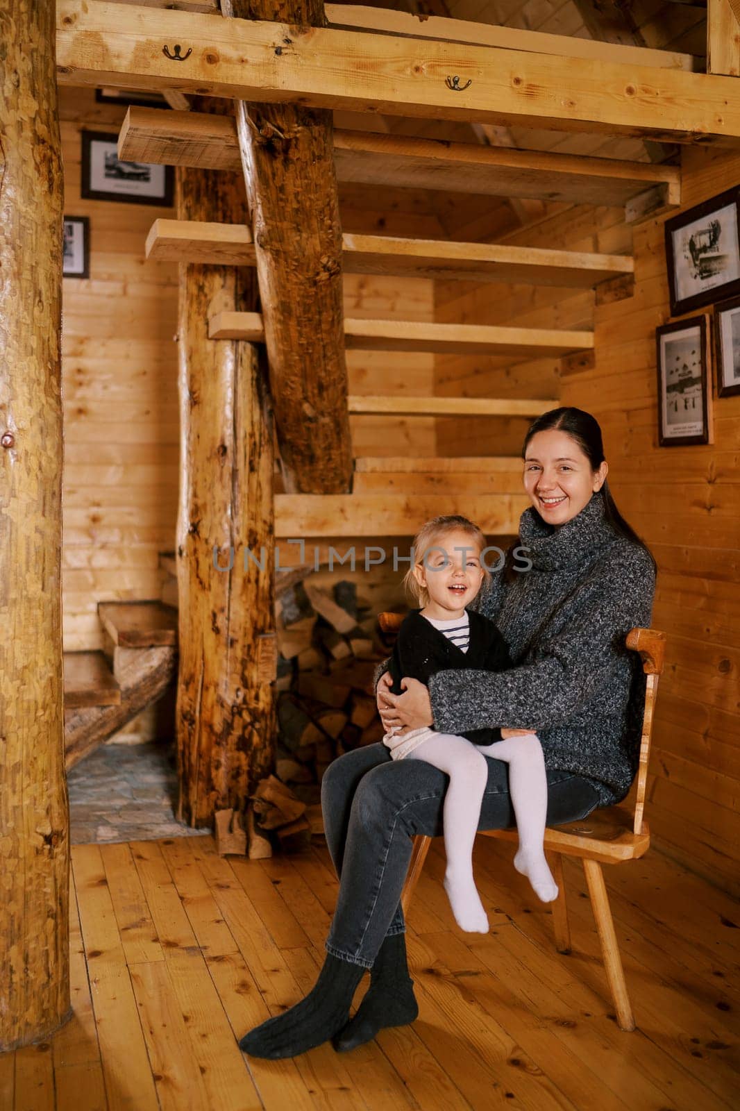 Smiling mother with little girl on lap sitting on chair near woodpile under stairs at home. High quality photo