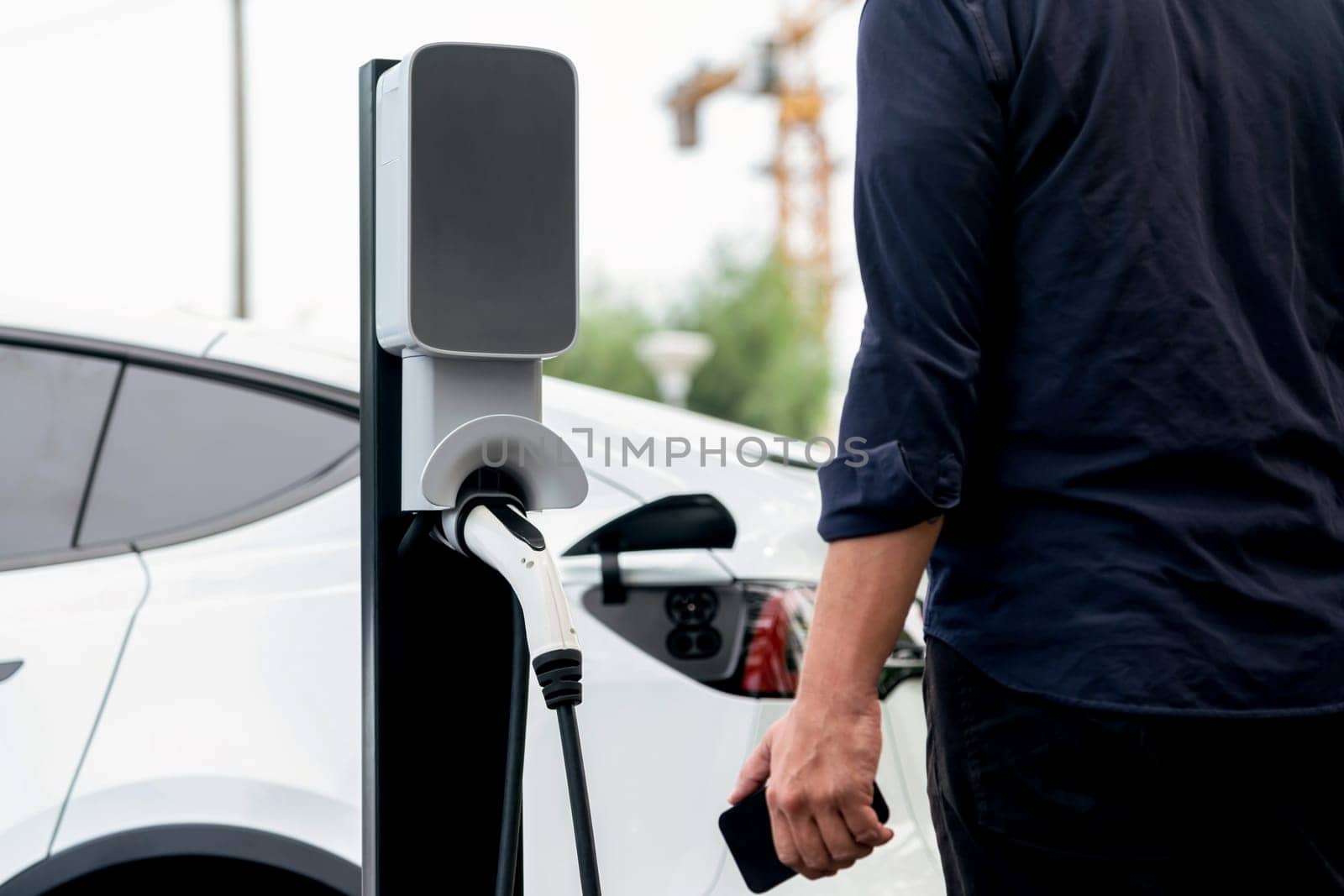 Man using smartphone online banking application to pay for electric car battery charging from EV charging station during vacation holiday road trip at national park or summer forest. Exalt