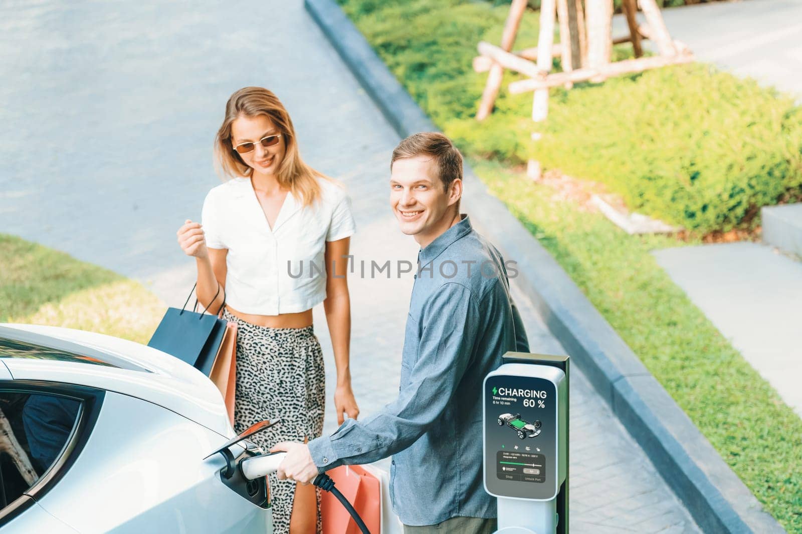 Young couple travel with EV electric car charging in green sustainable city outdoor garden in summer shows urban sustainability lifestyle by green clean rechargeable energy of electric vehicle innards