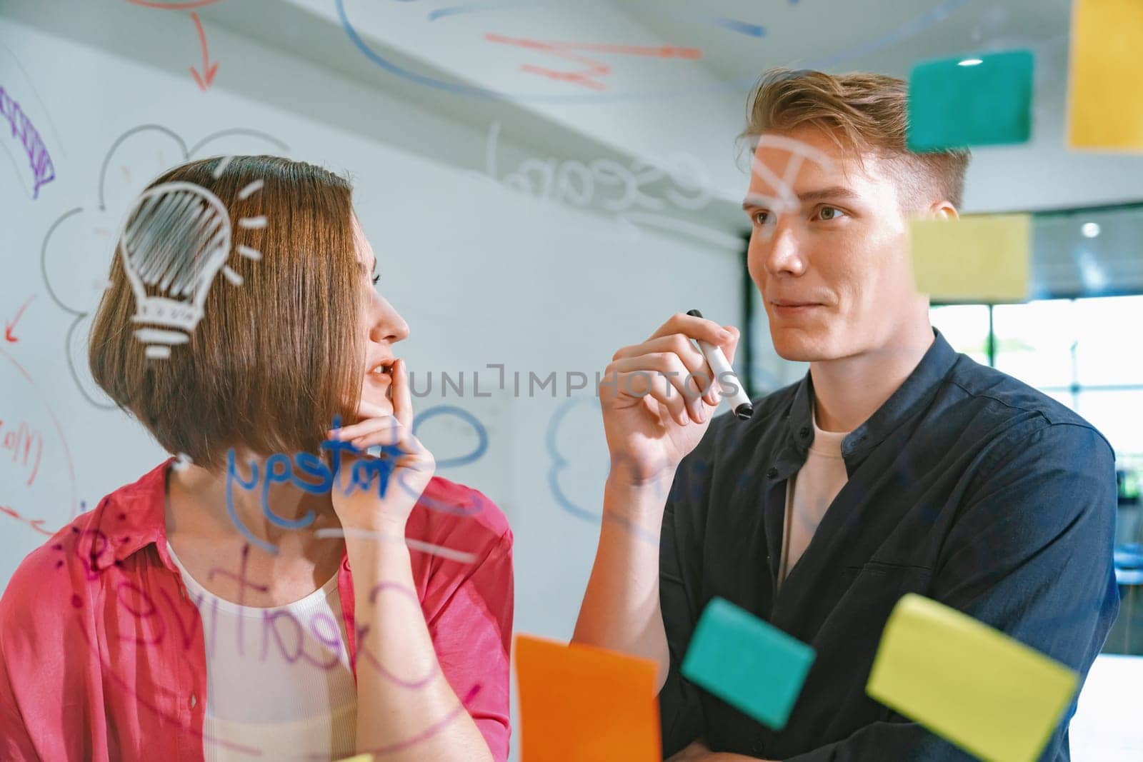 Couple of businesspeople discussing and brainstorming about solving marketing problem in front of glass board at business meeting by using mind map and sticky notes. Working together. Immaculate.