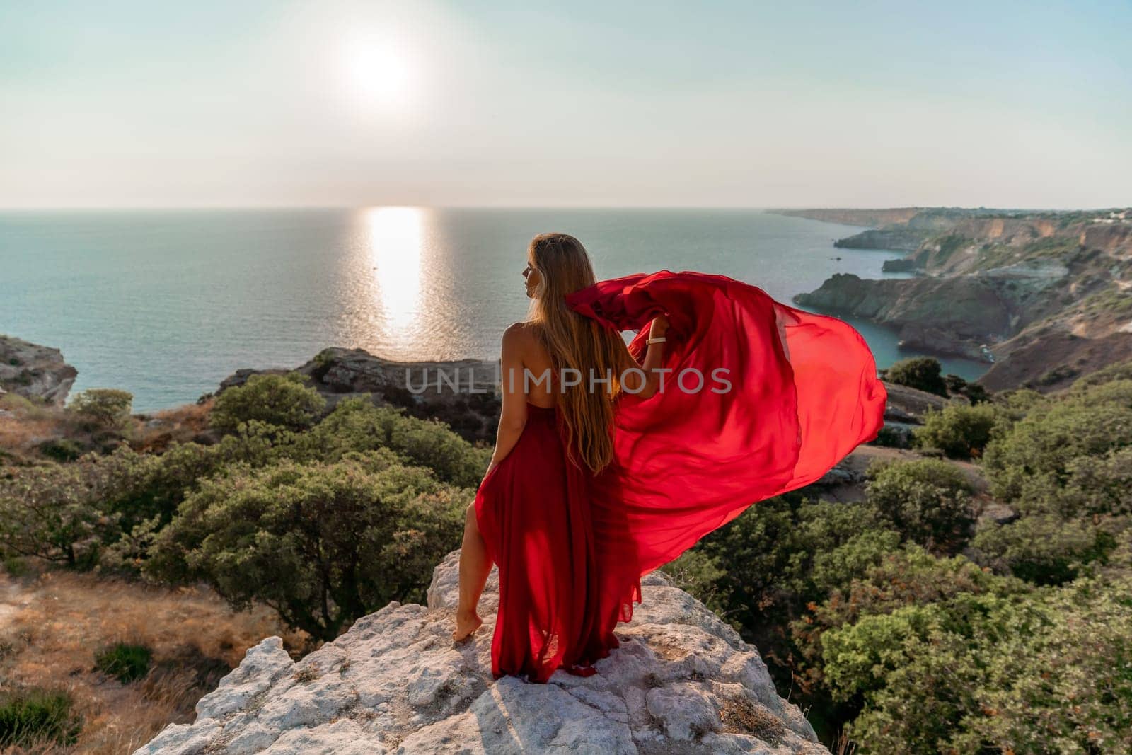 Woman sunset sea red dress, back view a happy beautiful sensual woman in a red long dress posing on a rock high above the sea on sunset. by Matiunina