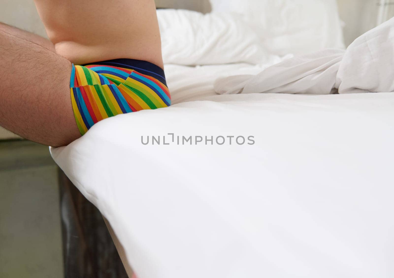 Unrecognizable gay man in rainbow pants, sitting on the white bedsheets on bed in the bedroom. Equality. Liberty. LGBT concept. Non binary person. Copy advertising space. Pride month