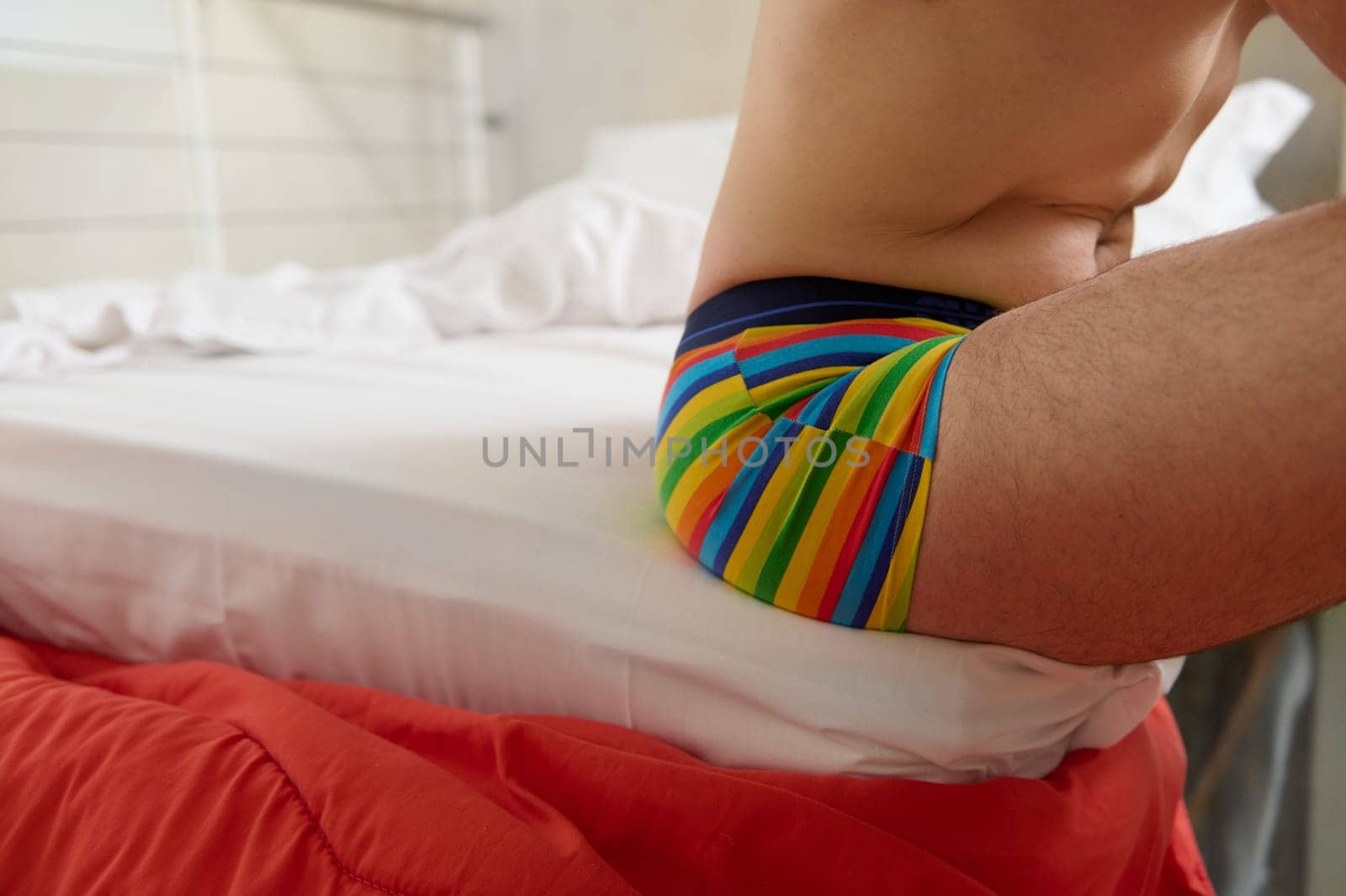 Unrecognizable young adult man of non-traditional orientation in rainbow underwear, sitting on the bed at home by artgf
