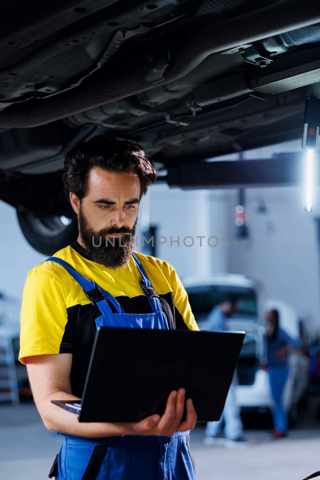 Diligent mechanic in garage using laptop to follow checklist while doing maintenance on car. Meticulous employee in auto repair shop does checkup on vehicle helped by device