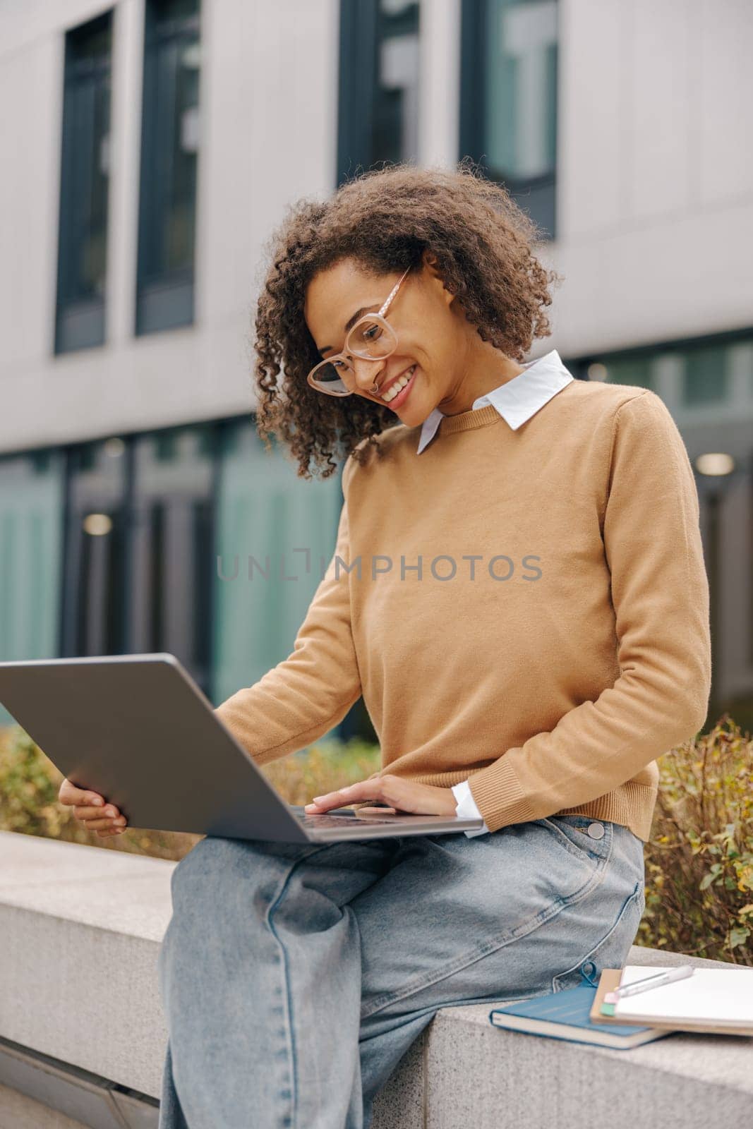 Smiling young female student working on laptop sitting on stairs on modern building background by Yaroslav_astakhov