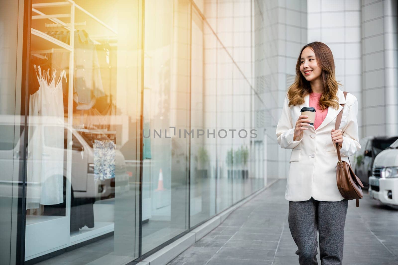 Happy successful Asian Businesswoman holding a coffee cup and brown bag on the Street next to dressing room a glass building. Happy girl finish work business lifestyle concept.