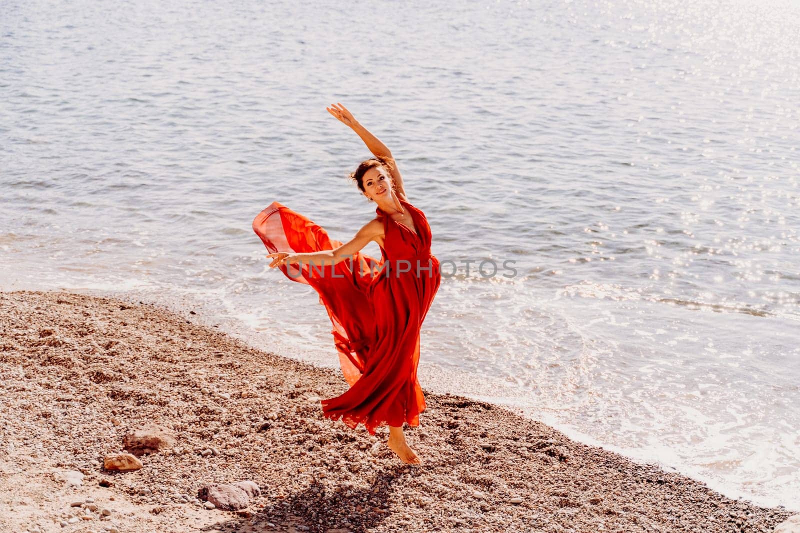 Woman red dress sea. Female dancer in a long red dress posing on a beach with rocks on sunny day. Girl on the nature on blue sky background