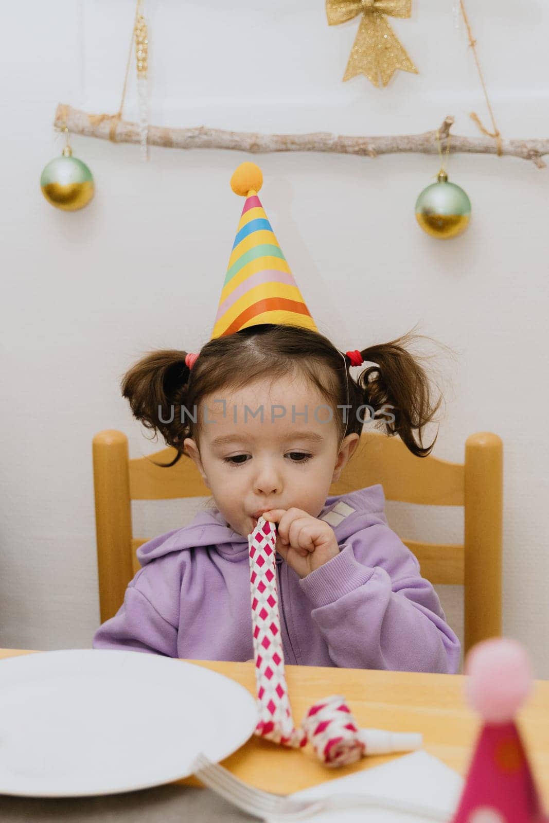 One beautiful Caucasian girl with a cone hat on her head sits at a festive table and blows a whistle into a paper pipe while celebrating her birthday, close-up side view.