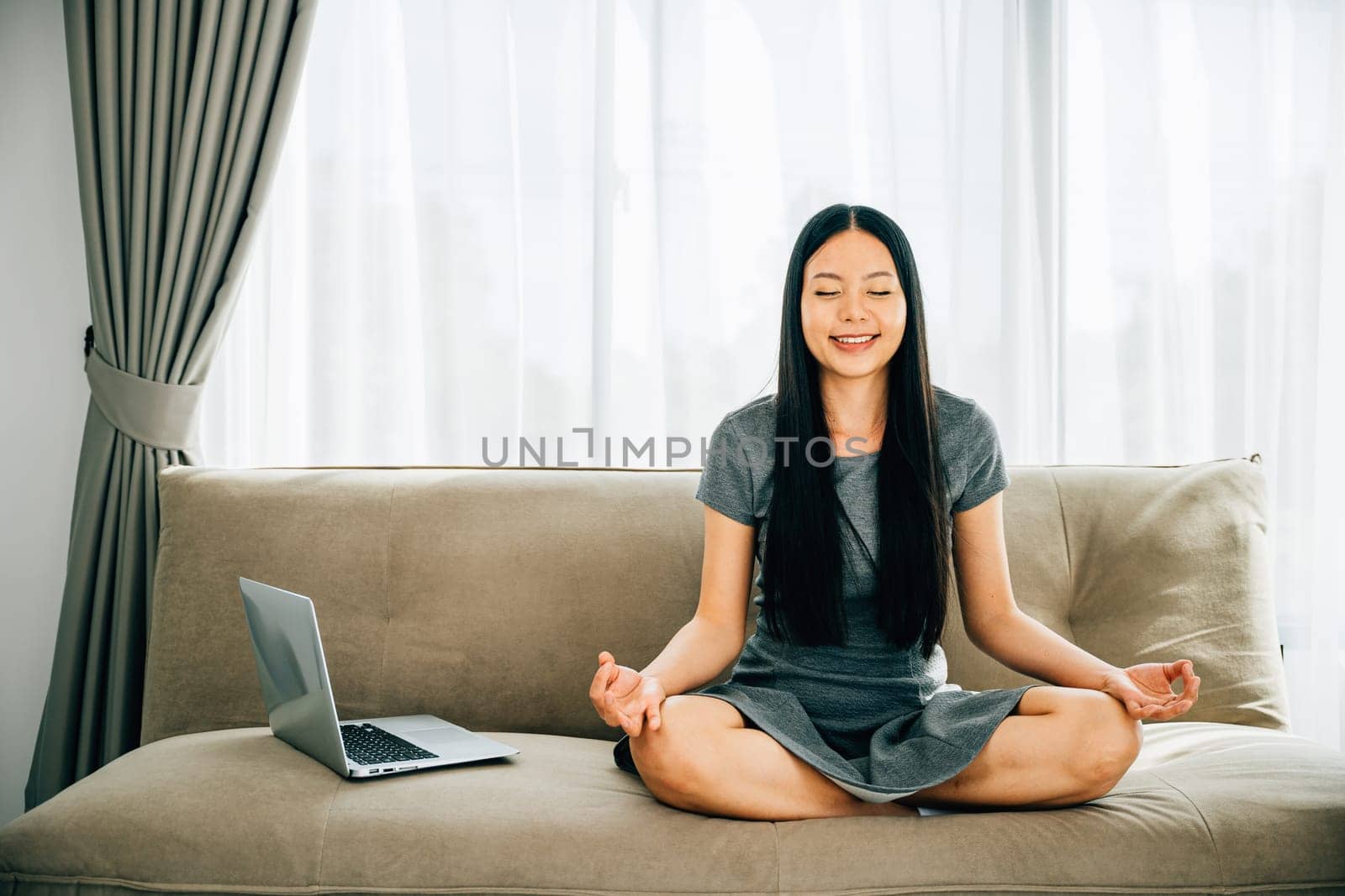 A serene Asian woman on sofa uses laptop meditating in lotus pose. Businesswoman finds relaxation and balance while practicing mindfulness online. Smiling student embodies harmony in the office.