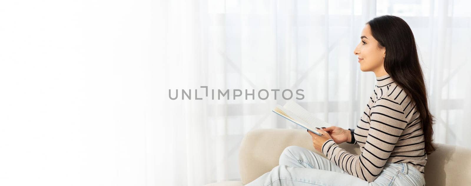 Smiling pensive calm young caucasian lady read book, planning work, sit at sofa, dream in light living room interior. Free time, study, education, hobby and lifestyle cozy at home