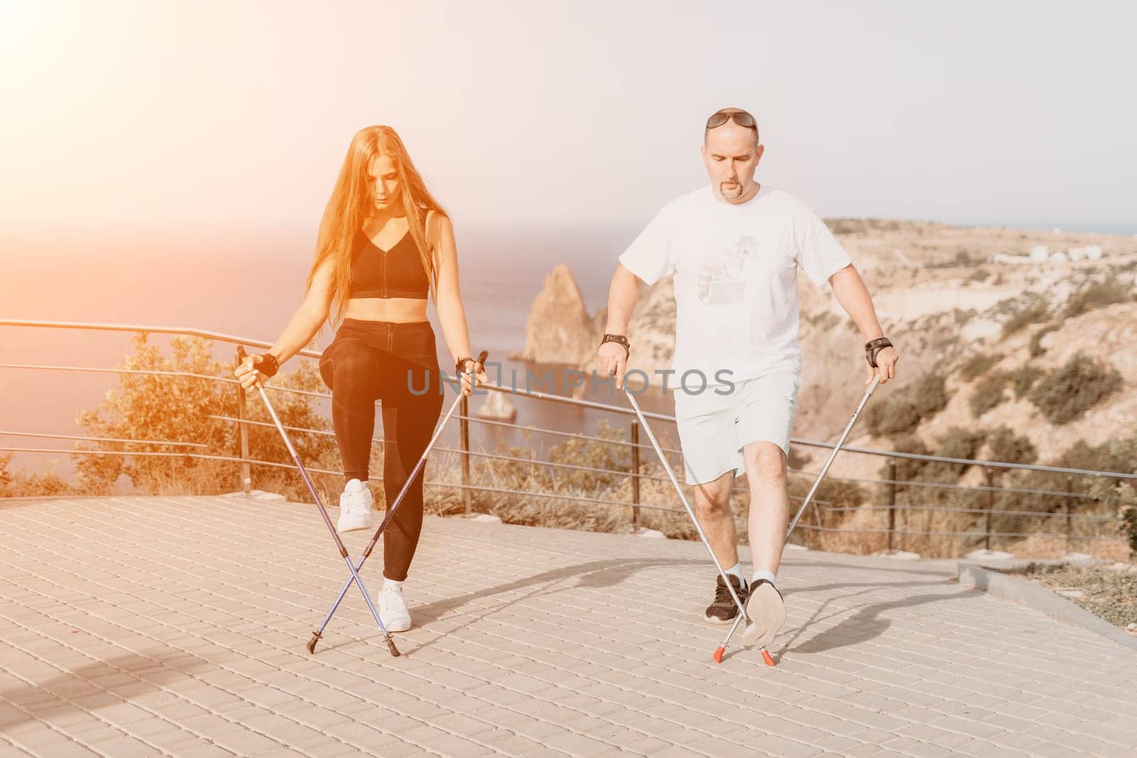 Happy Middle aged couple or friends practicing nordic walking in park near sea. Mature couple with trekking poles walking, practicing Nordic walking outdoors. Aging youthfully and sport concept.