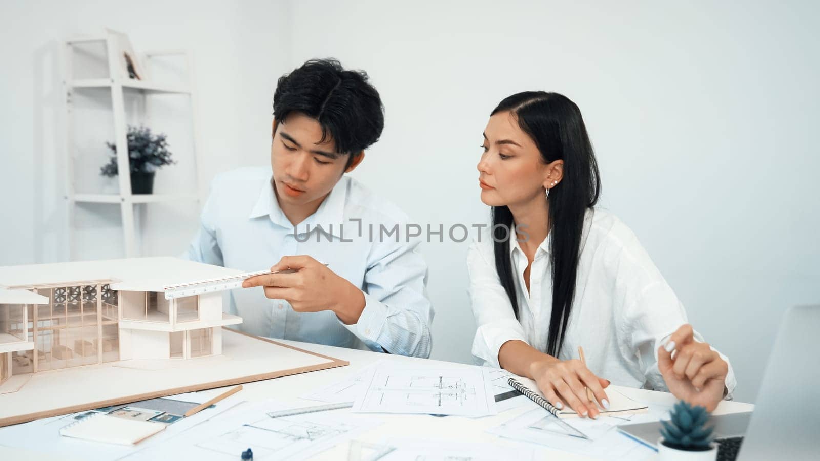 Professional male engineer measure house model by using ruler while beautiful cooperative coworker working together by using laptop to analysis data. Creative design and teamwork. Closeup. Immaculate.