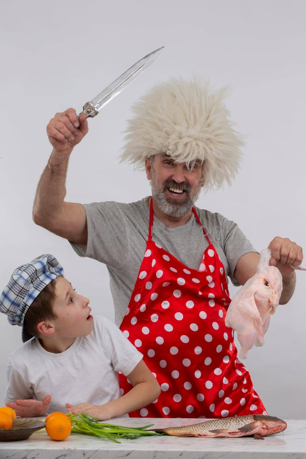 Georgian cheerful grandfather in a papakha hat teaches his grandson how to cook chicken. Master class in cooking. by Sviatlana