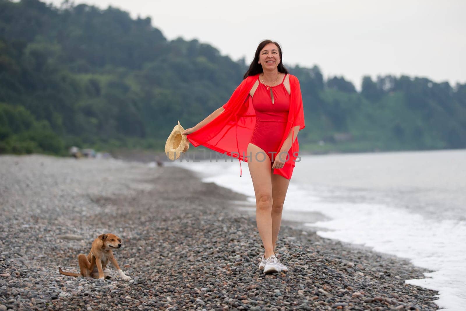 An elderly woman walks her dog along a pebbly beach.