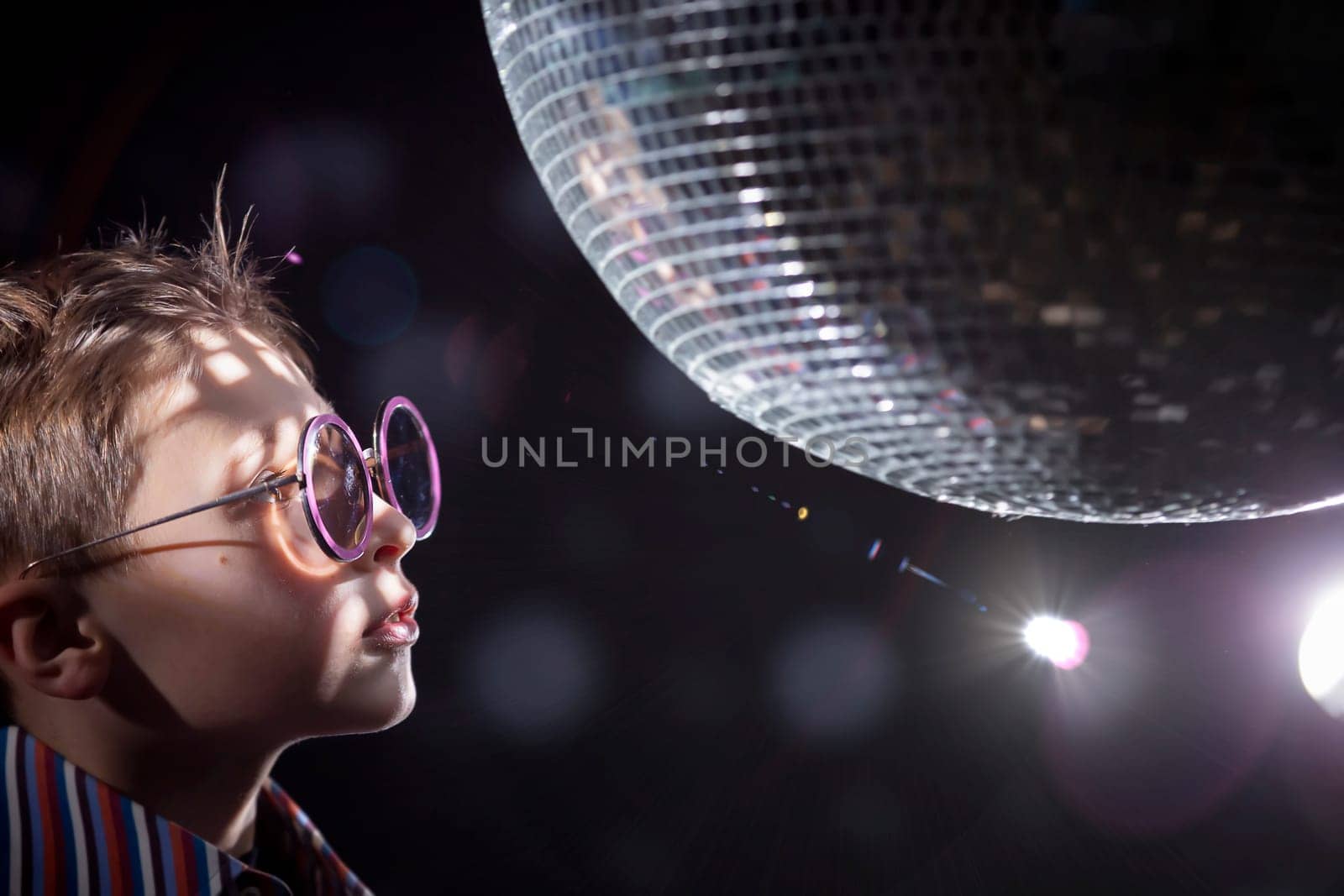 A young boy with glasses looks at a spinning disco ball. Children's disco, party.