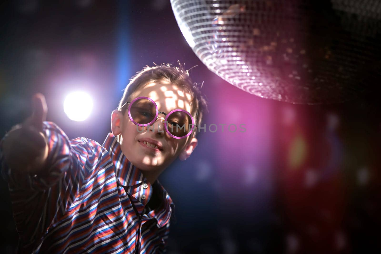 A young boy in stylish glasses shows a class sign against the background of light rays from a disco ball. Children's disco, party. by Sviatlana
