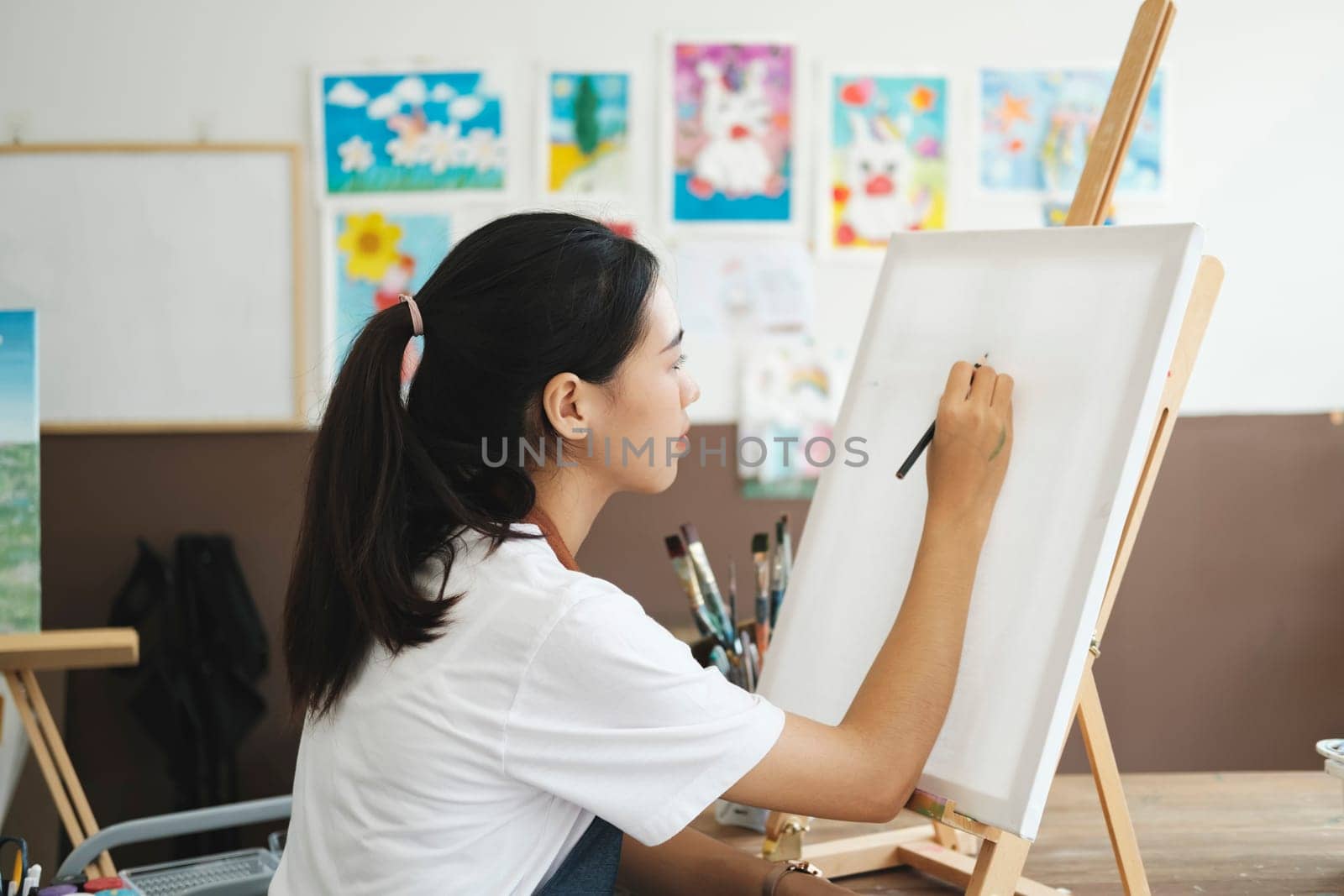 Young female artist sketches or paints her drawing on canvas in a studio workshop. A teenage girl who likes art and drawing is taking time to create her watercolors on canvas with great intention. Artist studio interior. Drawing supplies, oil paints, artist brushes, canvas, frame. Workshop or art class. Creative concept..