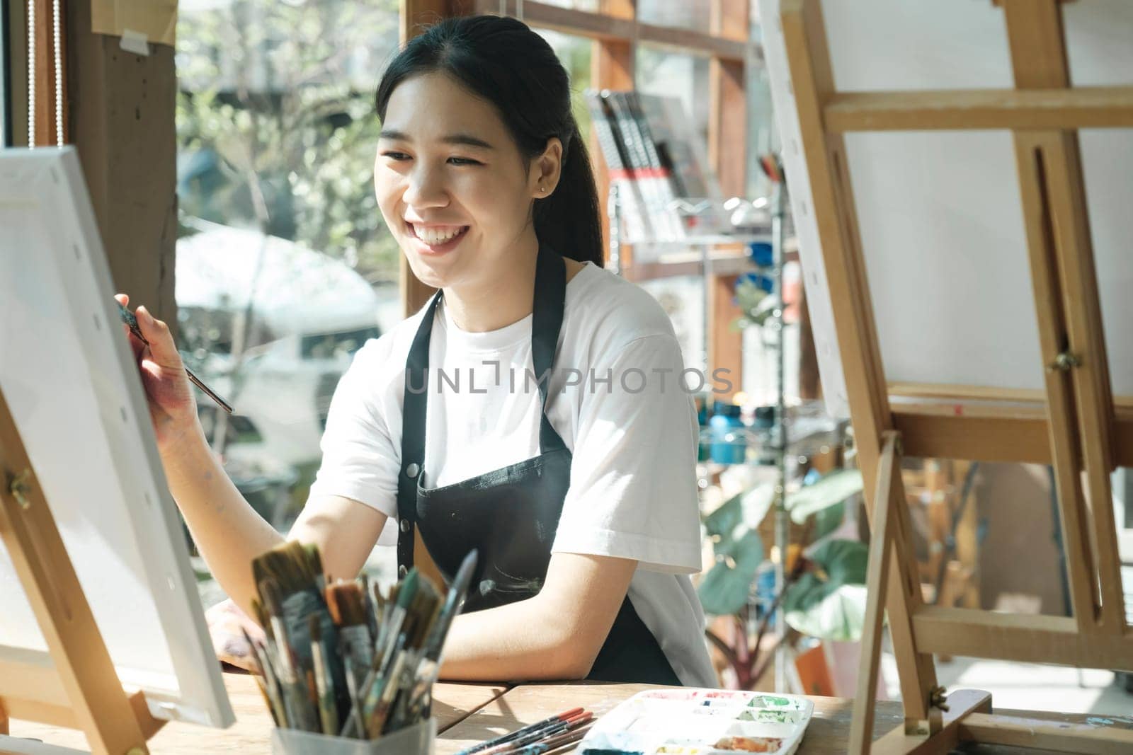 Young female artist sketches or paints her drawing on canvas in a studio workshop. A teenage girl who likes art and drawing is taking time to create her watercolors on canvas with great intention. Artist studio interior. Drawing supplies, oil paints, artist brushes, canvas, frame. Workshop or art class. Creative concept..