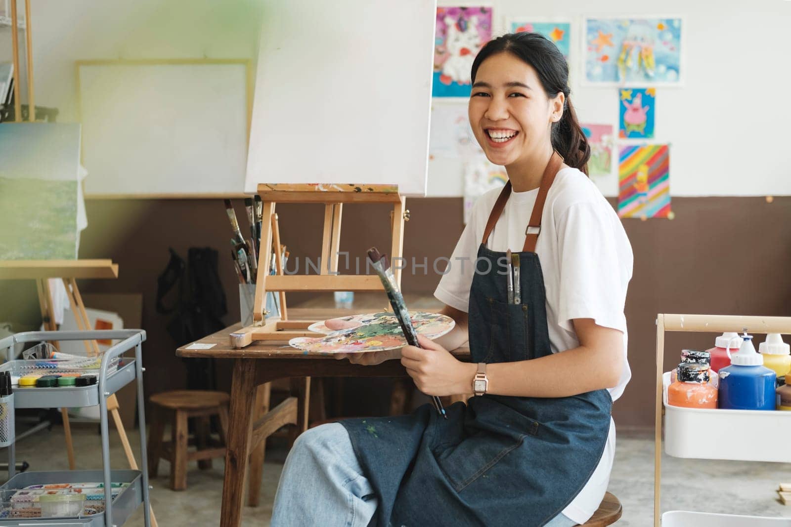 The young artist female is wearing an apron smeared with paint clutching many brushes and paintbrushes stnading and smiling to the camera in her studio workshop or art gallery. Hobby and lifestyle concept.