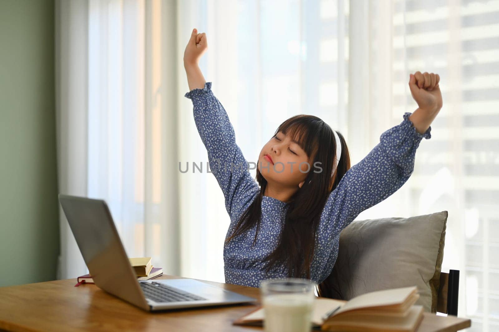 Tired schoolgirl stretching in front of laptop while studying remotely at home. Online education concept.