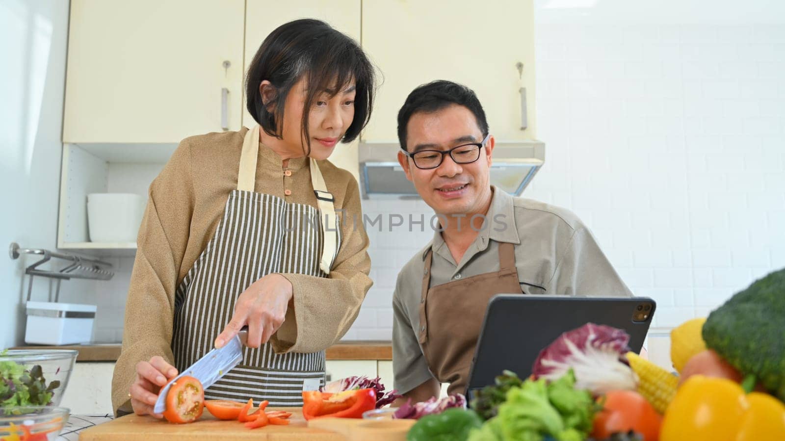 Senior couple reading recipe from internet on digital tablet while cooking healthy food together in kitchen by prathanchorruangsak