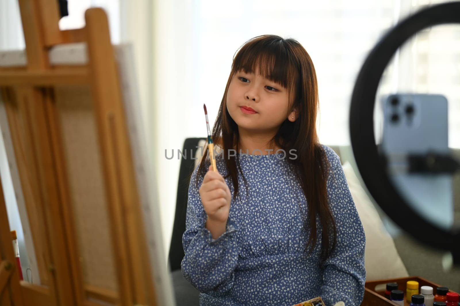 Cute Asian schoolgirl painting picture and recording video on smartphone at home. by prathanchorruangsak
