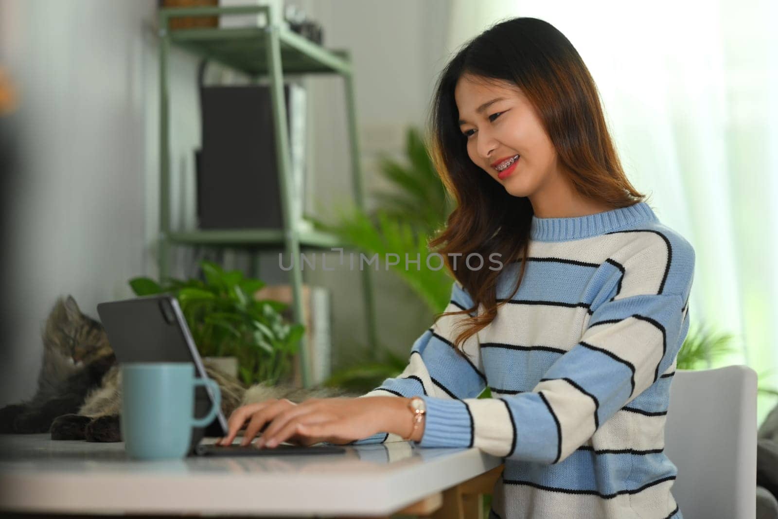 Beautiful young woman in warm sweater using laptop on white desk at cozy home.