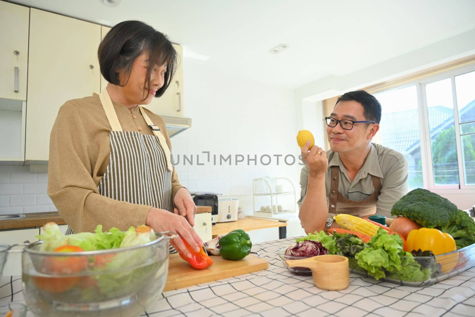 Loving Asian senior couple wearing aprons cooking together at home kitchen. Retirement lifestyle concept by prathanchorruangsak