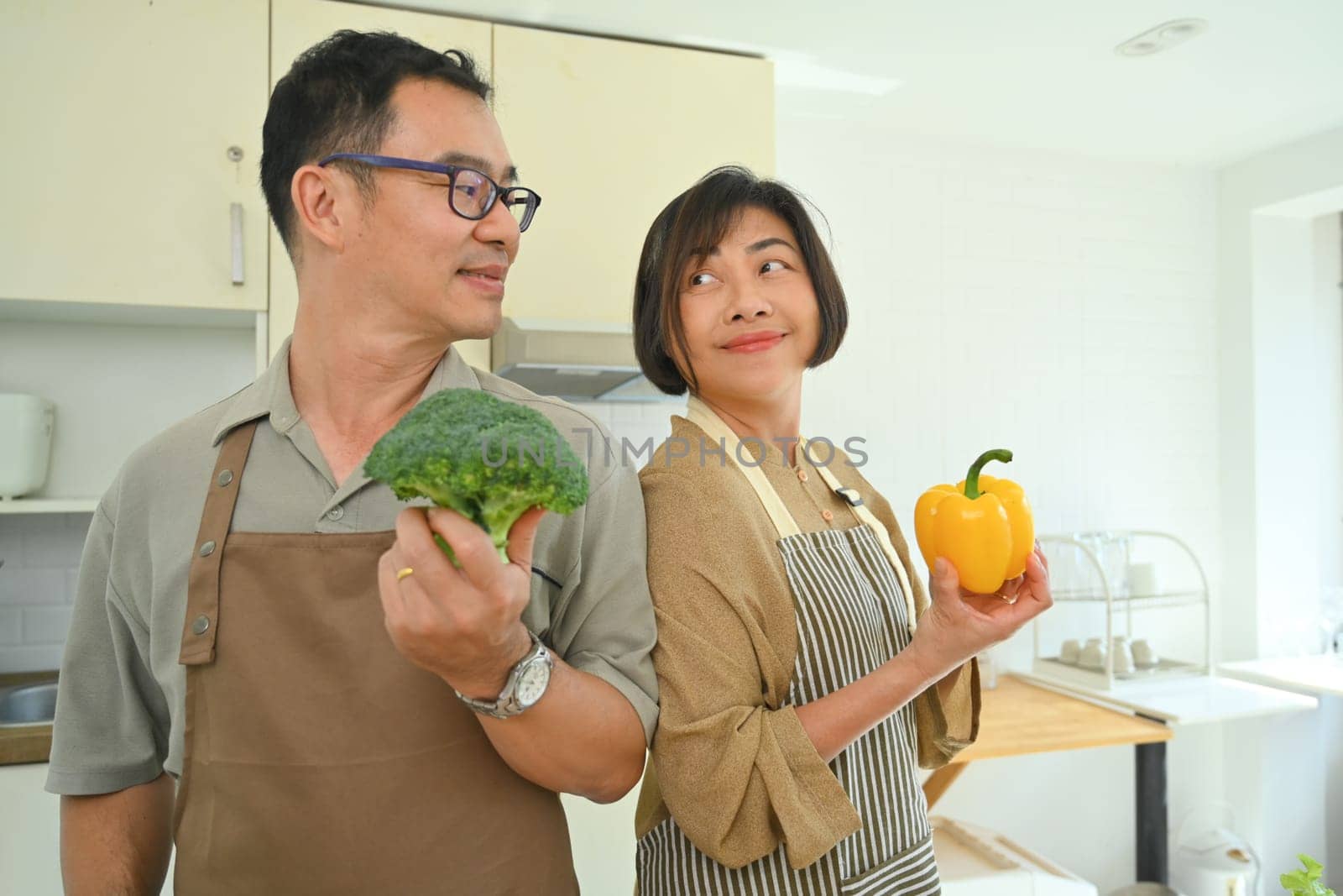 Happy older husband and wife in aprons preparing vegetarian meal together in kitchen by prathanchorruangsak