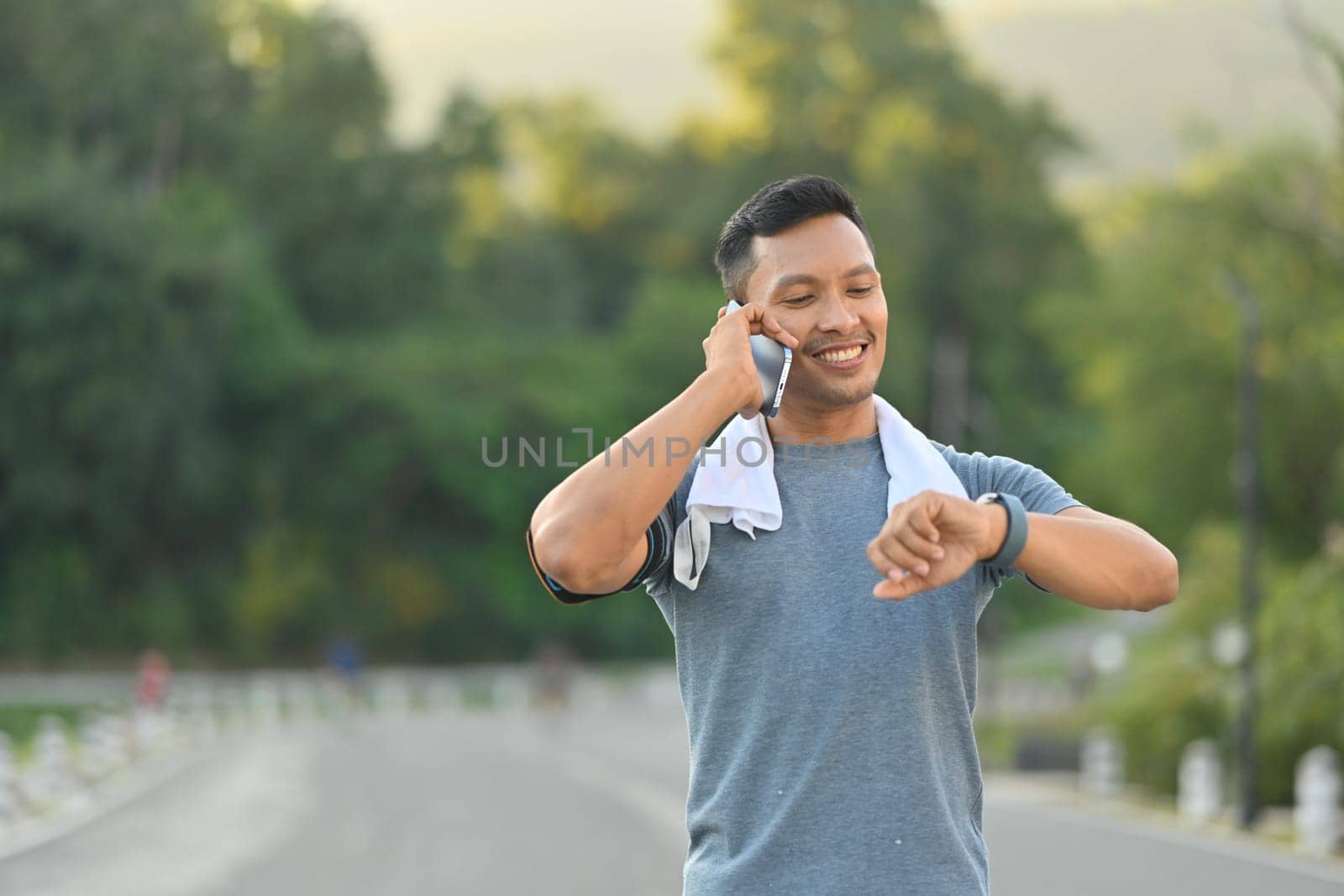 Smiling sportsman having pleasant phone conversation and checking time on watch while exercise in the park by prathanchorruangsak