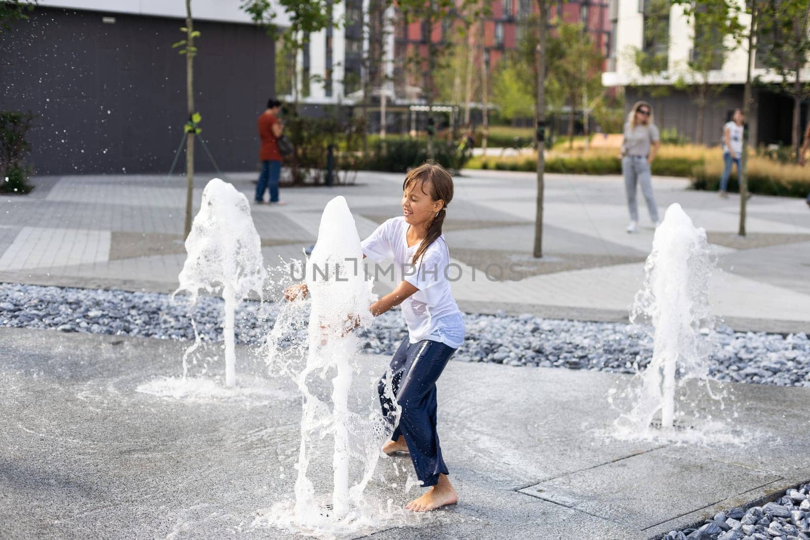 Cheerful young teen girl in city fountain, girl in wet clothes is having fun and enjoying the cool summer water, background city architecture. by Andelov13