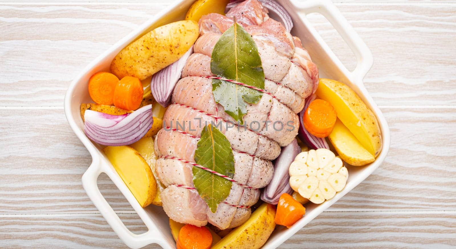 Raw wrapped rolled sliced pork in white casserole dish with potatoes, vegetables and herbs on rustic white wooden background top view. Pork roll with vegetables ready to be prepared.