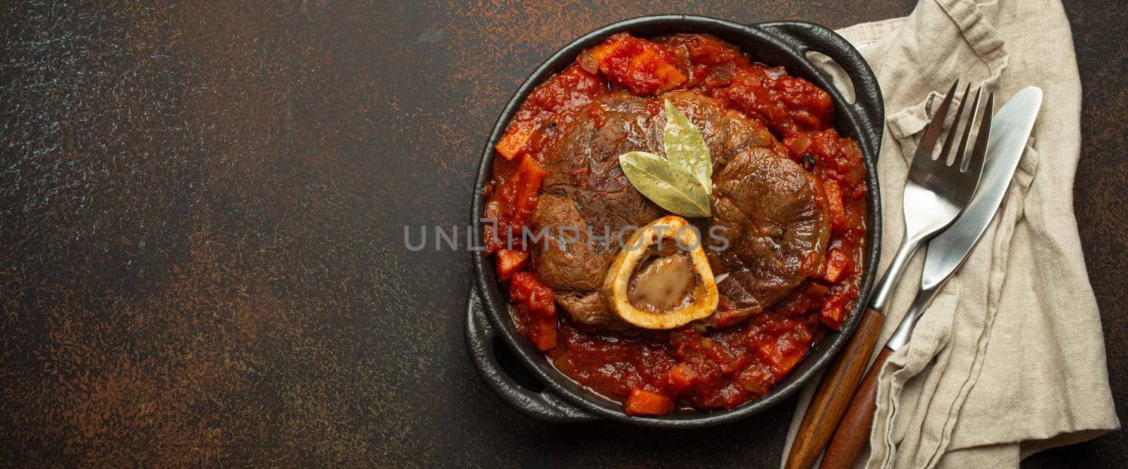 Traditional Italian dish Ossobuco all Milanese made with cut veal shank meat with vegetable tomato sauce served in black casserole pan top view on rustic brown background, copy space.