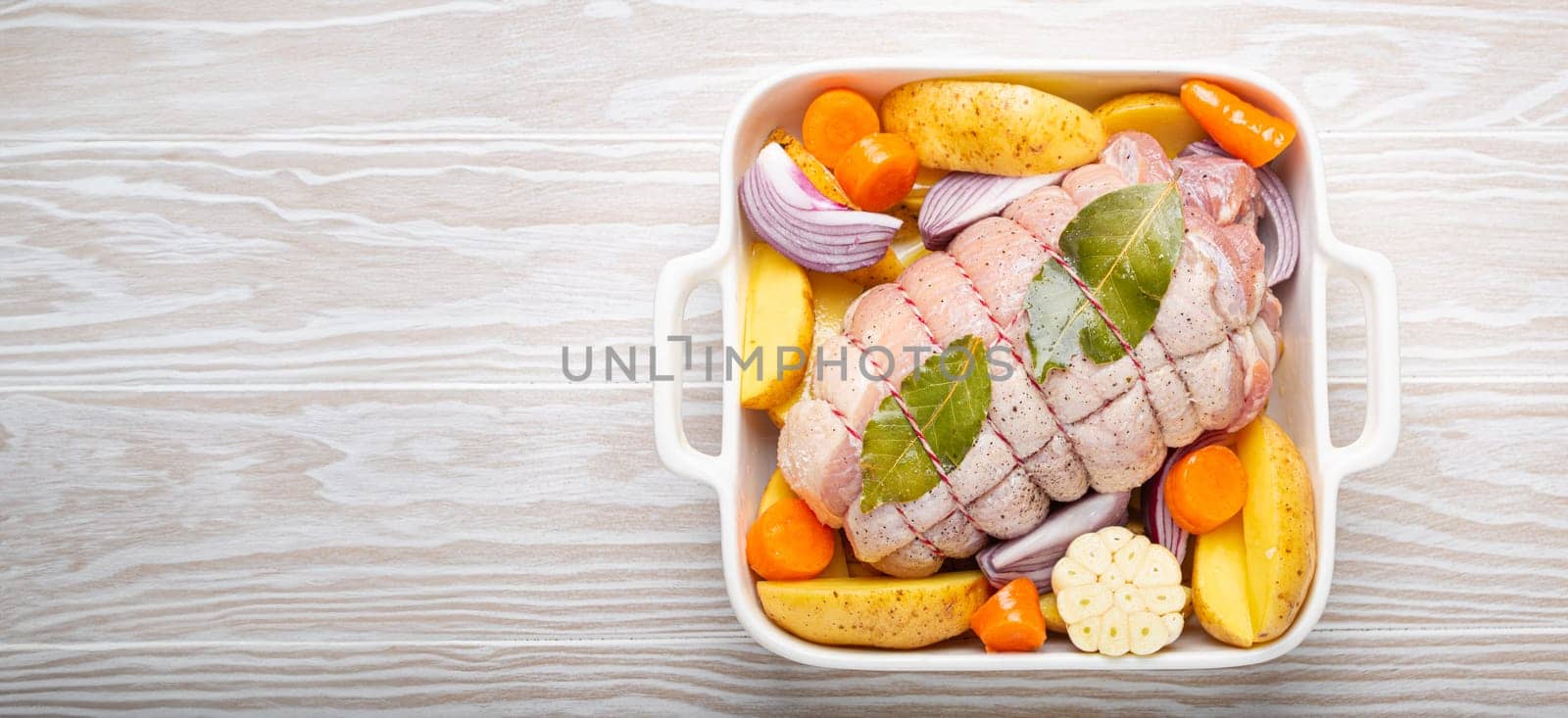 Raw wrapped rolled sliced pork in white casserole dish with potatoes, vegetables and herbs on rustic white wooden background top view. Pork roll with vegetables ready to be prepared.