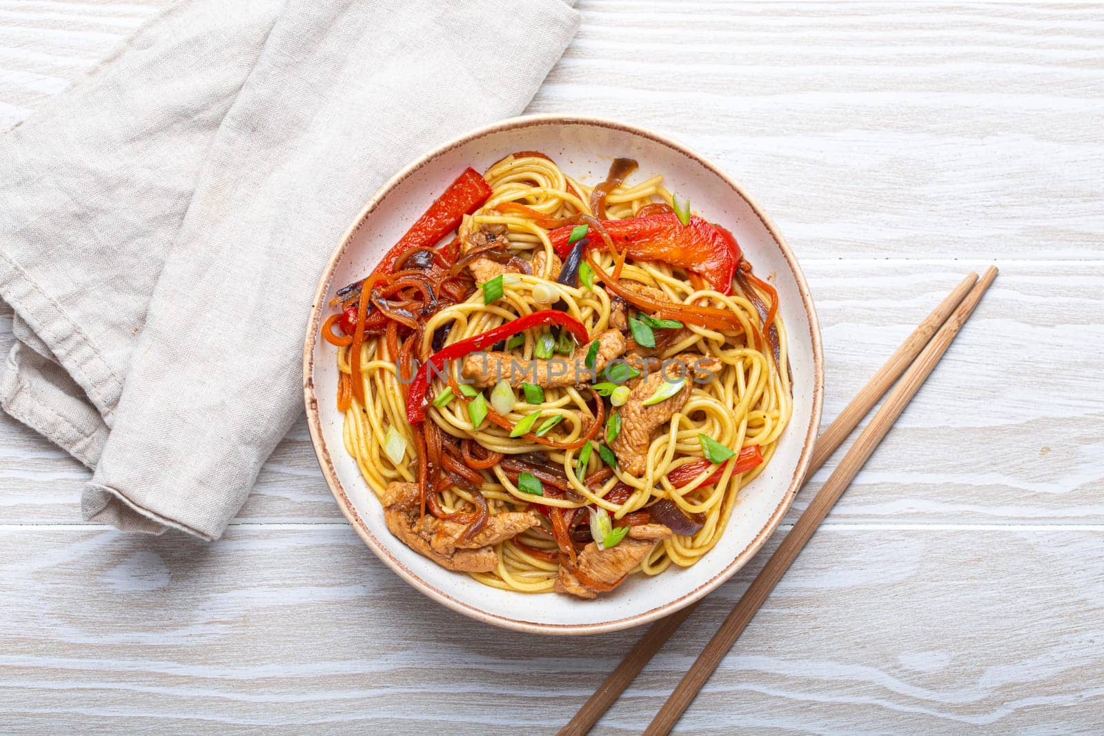 White bowl with Chow Mein or Lo Mein, traditional Chinese stir fry noodles with meat and vegetables, served with chopsticks top view on rustic white wooden background table.