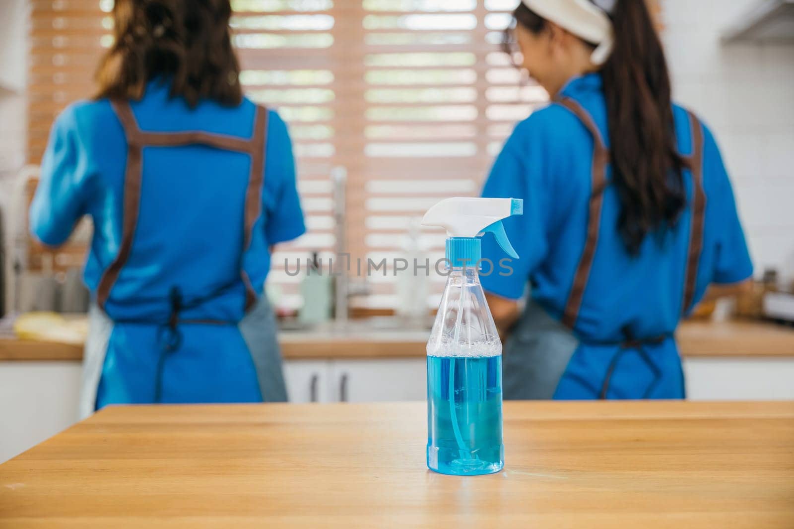 Occupied woman cleaning kitchen desk using spray product and towel. Housekeeping concept with a focus on hygiene and safety. Uniformed cleaner at work. Clean disinfect home care. Closeup spray bottle. by Sorapop