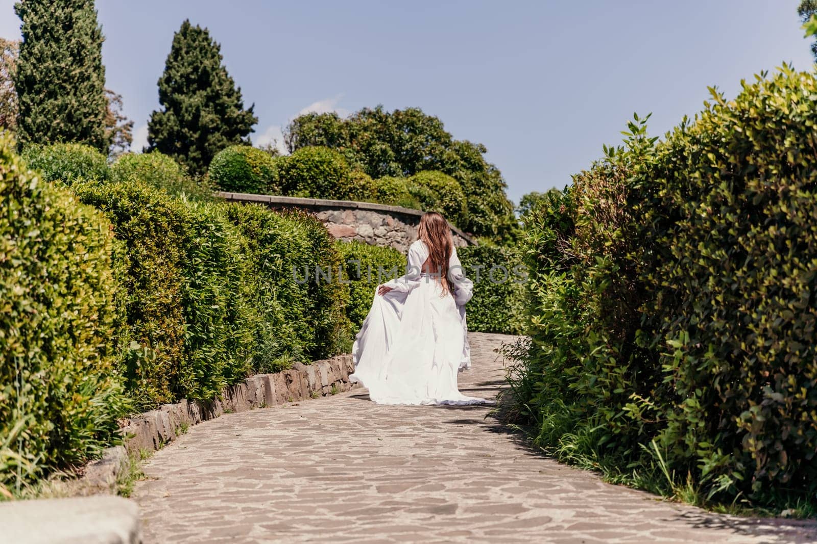 Brunette runs white dress park. A beautiful woman with long brown hair and a long white dress runs along the path along the beautiful bushes in the park, rear view by Matiunina