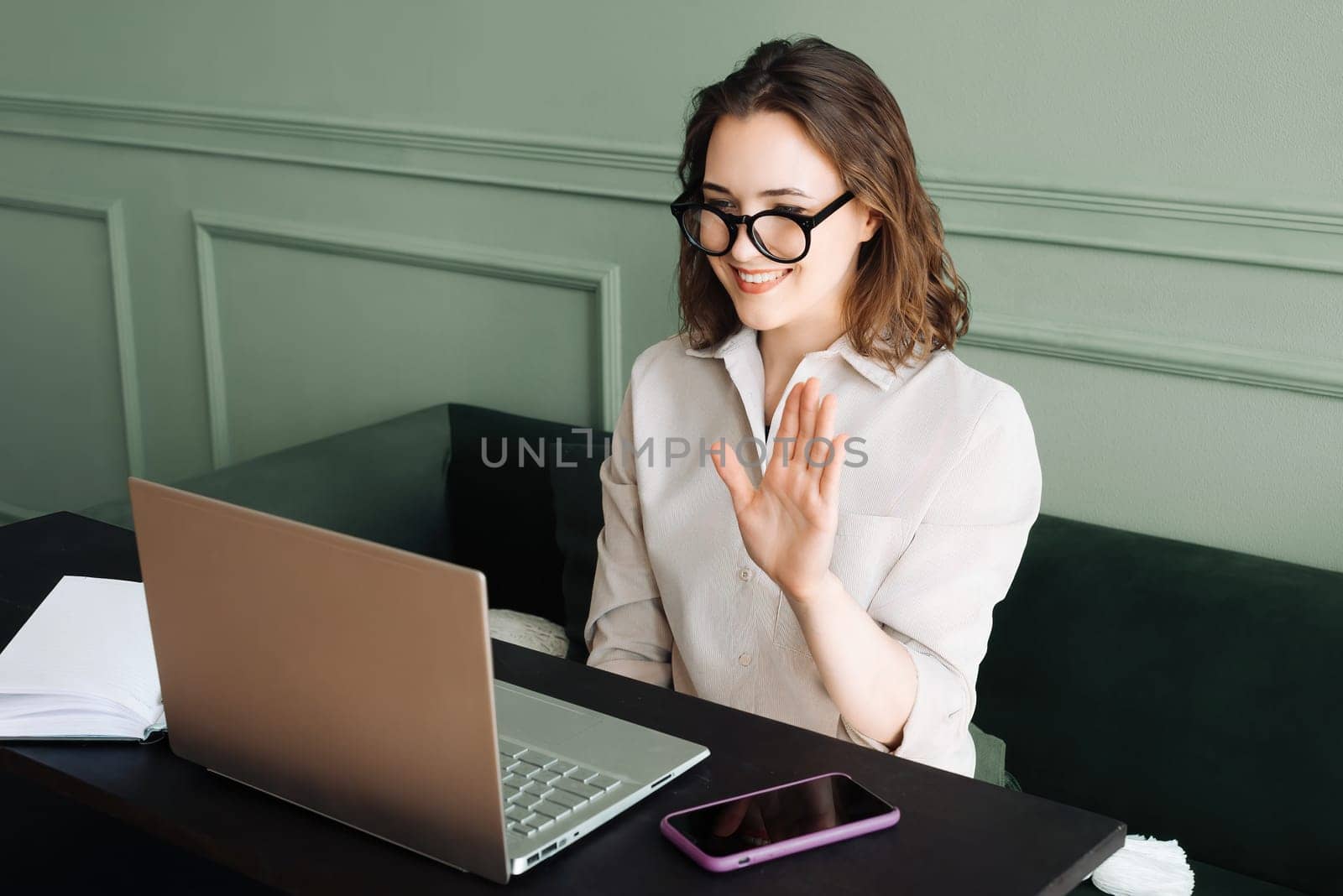 Cheerful Video Chat. Young Woman with Glasses Sharing Greetings. Expressive Online Interaction: Joyful Woman Waves in Laptop Call.