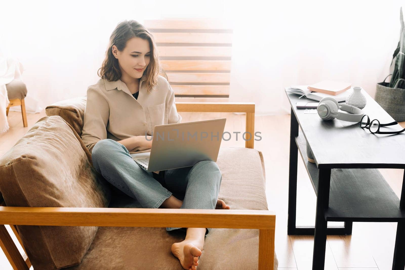 Joyful millennial woman typing on a laptop, attending a virtual meeting, seated on a sofa. Cheerful millennial female typing on laptop, has meeting with gadget, sit on sofa.