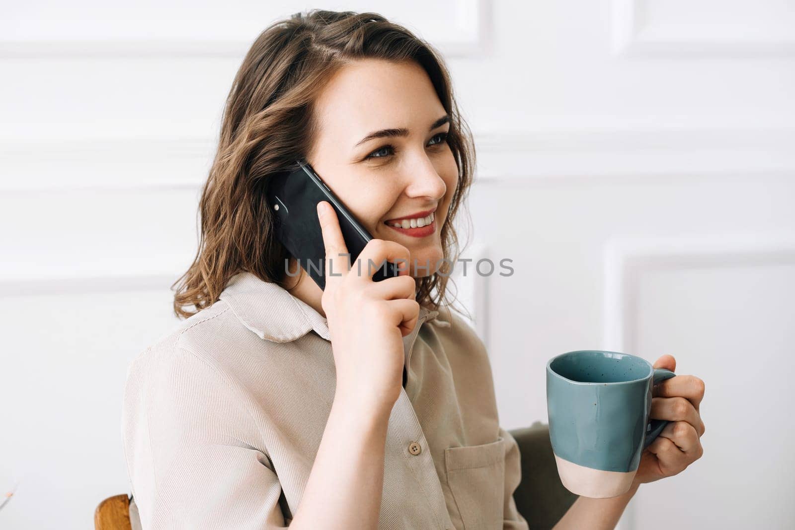 Happy Young Woman Enjoying a Phone Call While Savoring a Drink in Leisure Time. Joyful Young Woman Talking on Phone, Enjoying a Relaxing Moment with a Cup of Drink in Her Free Time.