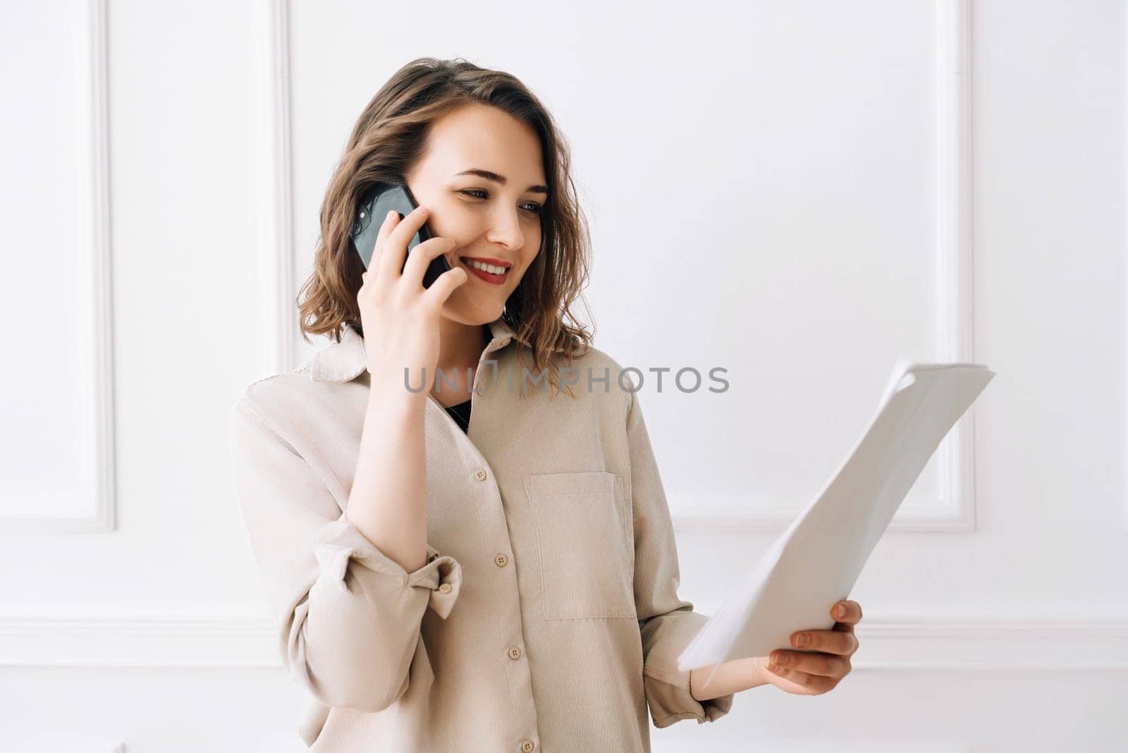 Smiling Millennial Woman Engaged in Phone Call, Discussing Documents with Client. Smiling millennial woman calling by smartphone, talking with client, looking at documents.