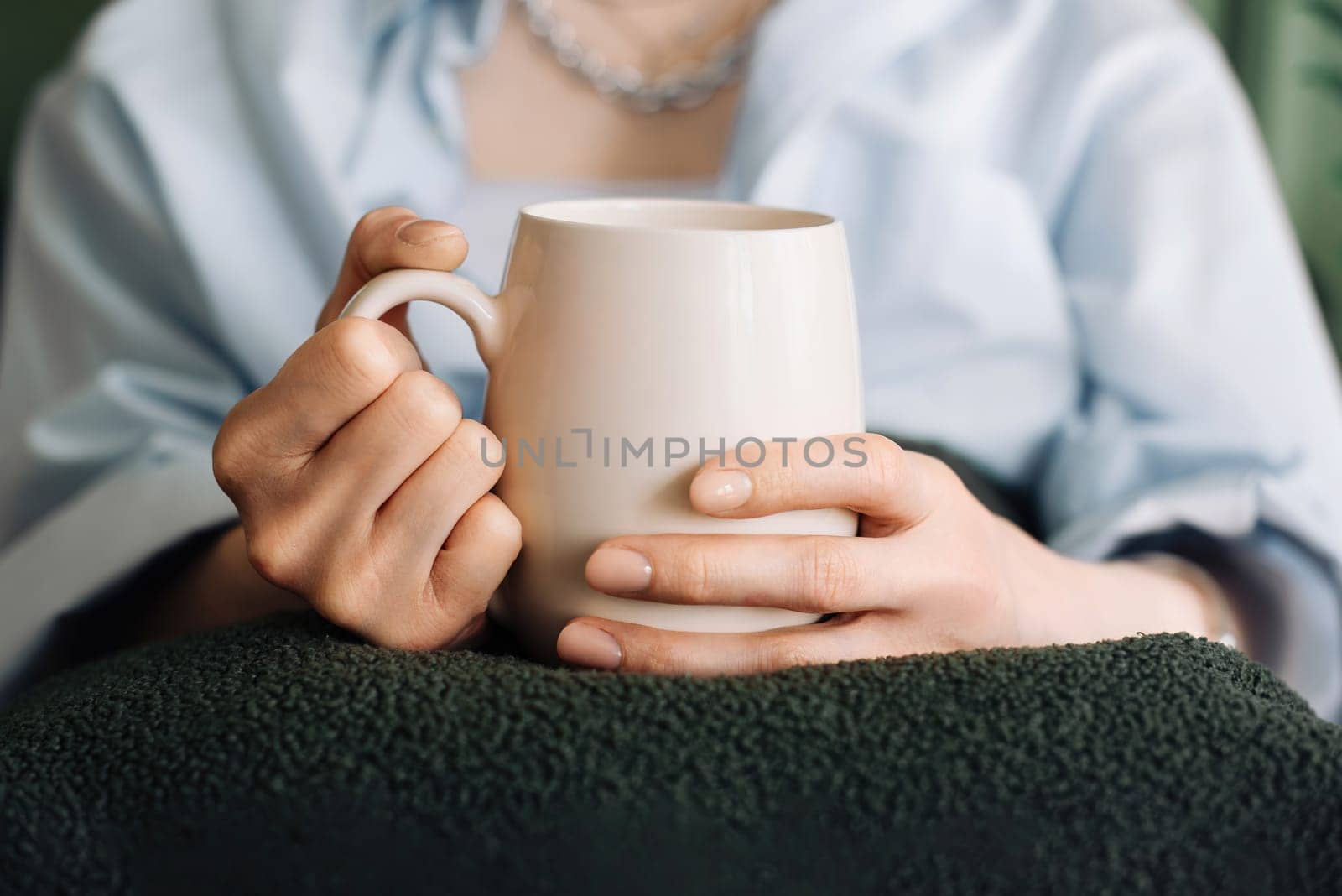 Woman Enjoying Tea on Living Room Couch, Cropped View. Cozy Domestic Scene. Young Woman Unwinding with Tea in Living Room Interior. Female hands holding a mug with tea or coffee. Close-up.