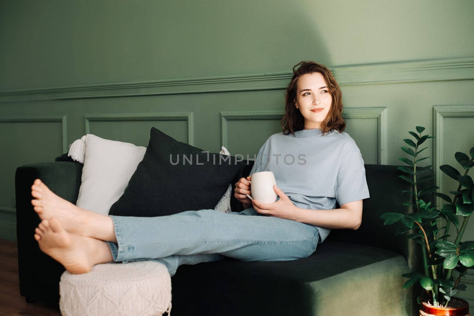 A thoughtful young middle-aged woman enjoying a rest and a cup of tea on the couch, pondering, looking at the open space. Pensive Moment. Thoughtful Young Middle-aged Woman Rests with Tea on Couch.