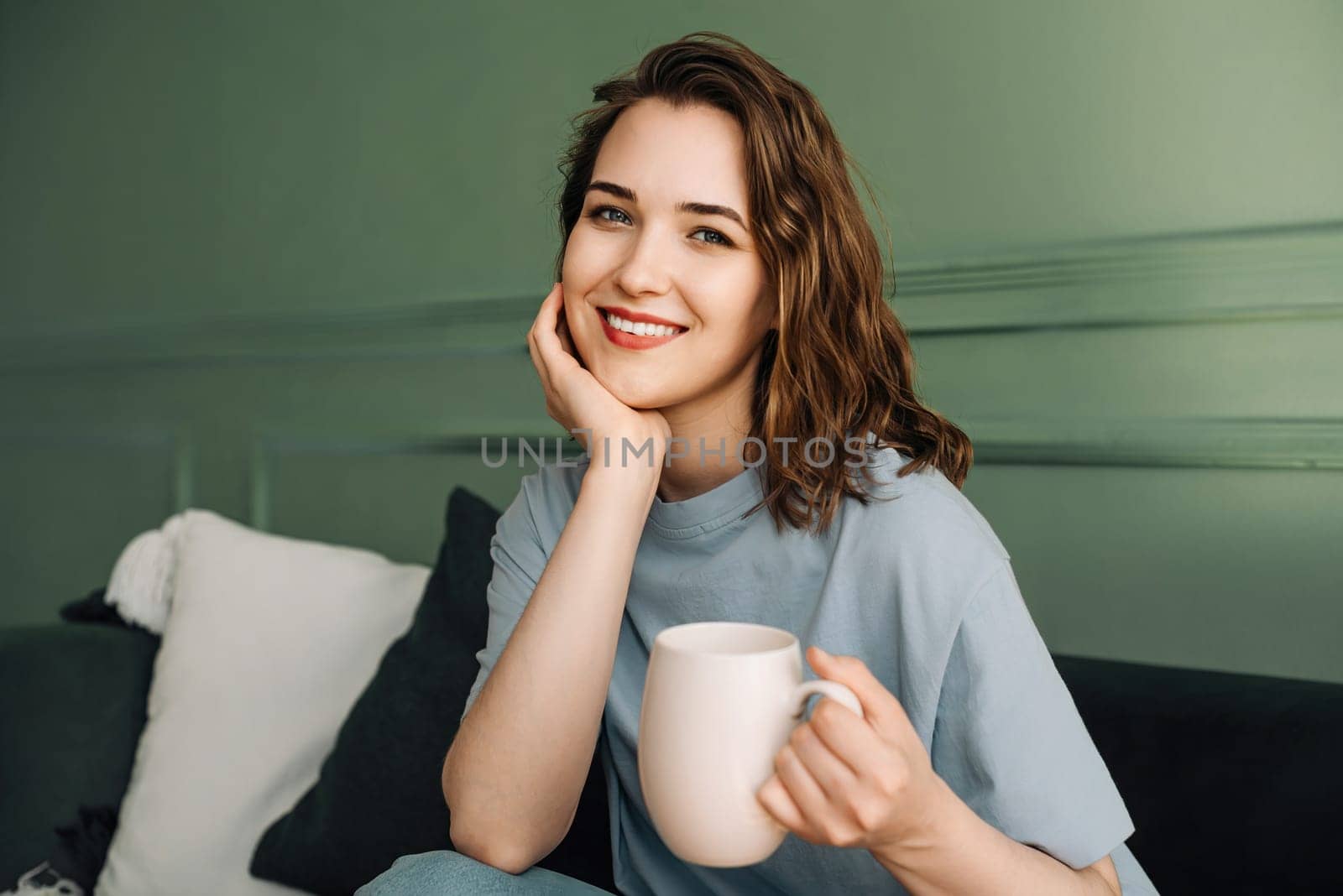 Relaxed Alone Time. Smiling Woman Enjoying Hot Drink on Couch in Living Room. Cozy Leisure Moment: Young Woman Savoring Drink on Sofa, Smiling in Living Room Ambiance