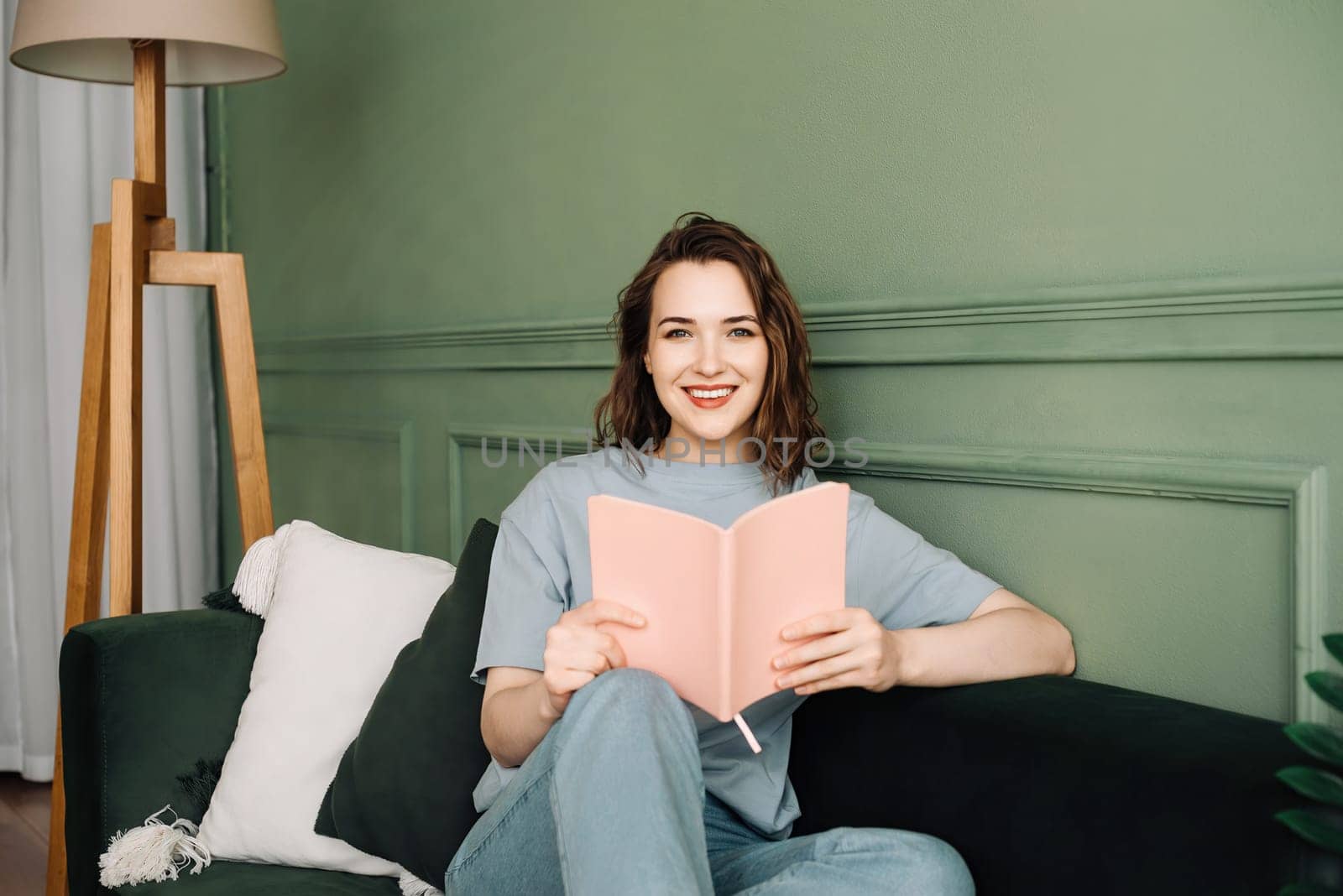 Blissful Young Woman Engaged in Reading, Relaxing on Couch During Serene Evening. Content Young Woman Immersed in Book, Relaxed on Couch in Pleasant Evening Ambiance.