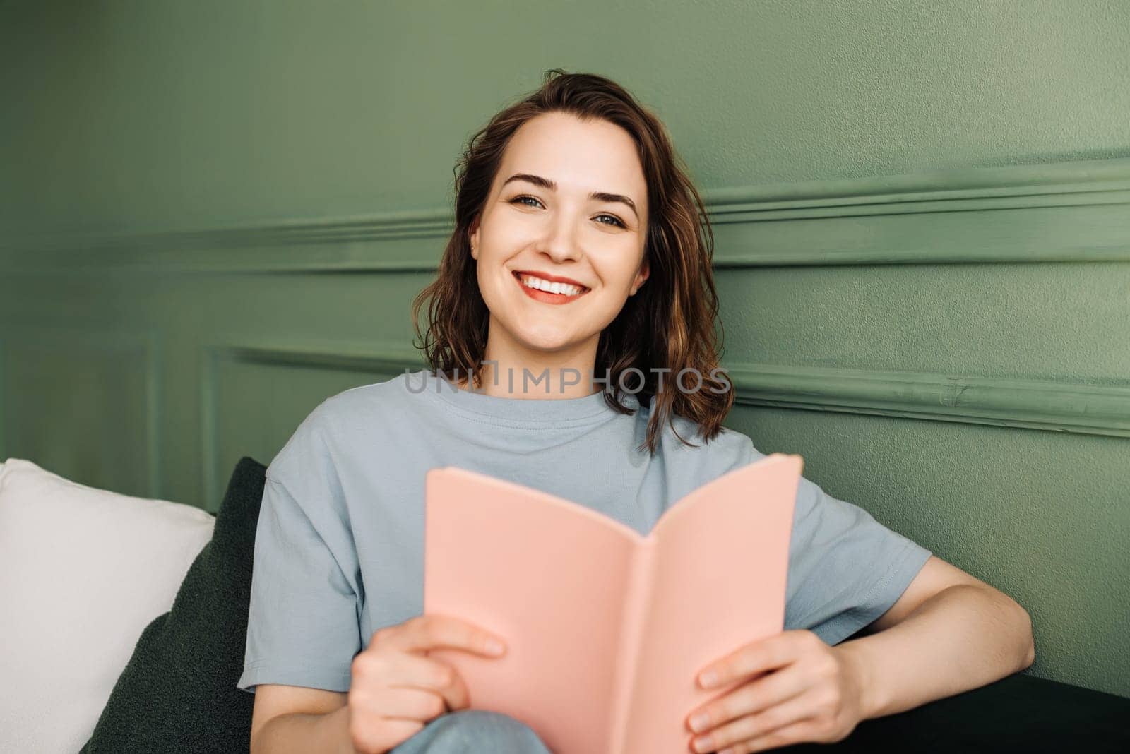 Content Young Woman Reading Book on Couch. Relaxed Girl Enjoying Evening Reading on Sofa. Leisure Time and Relaxation: Happy Woman Reading Book Comfortably on Couch