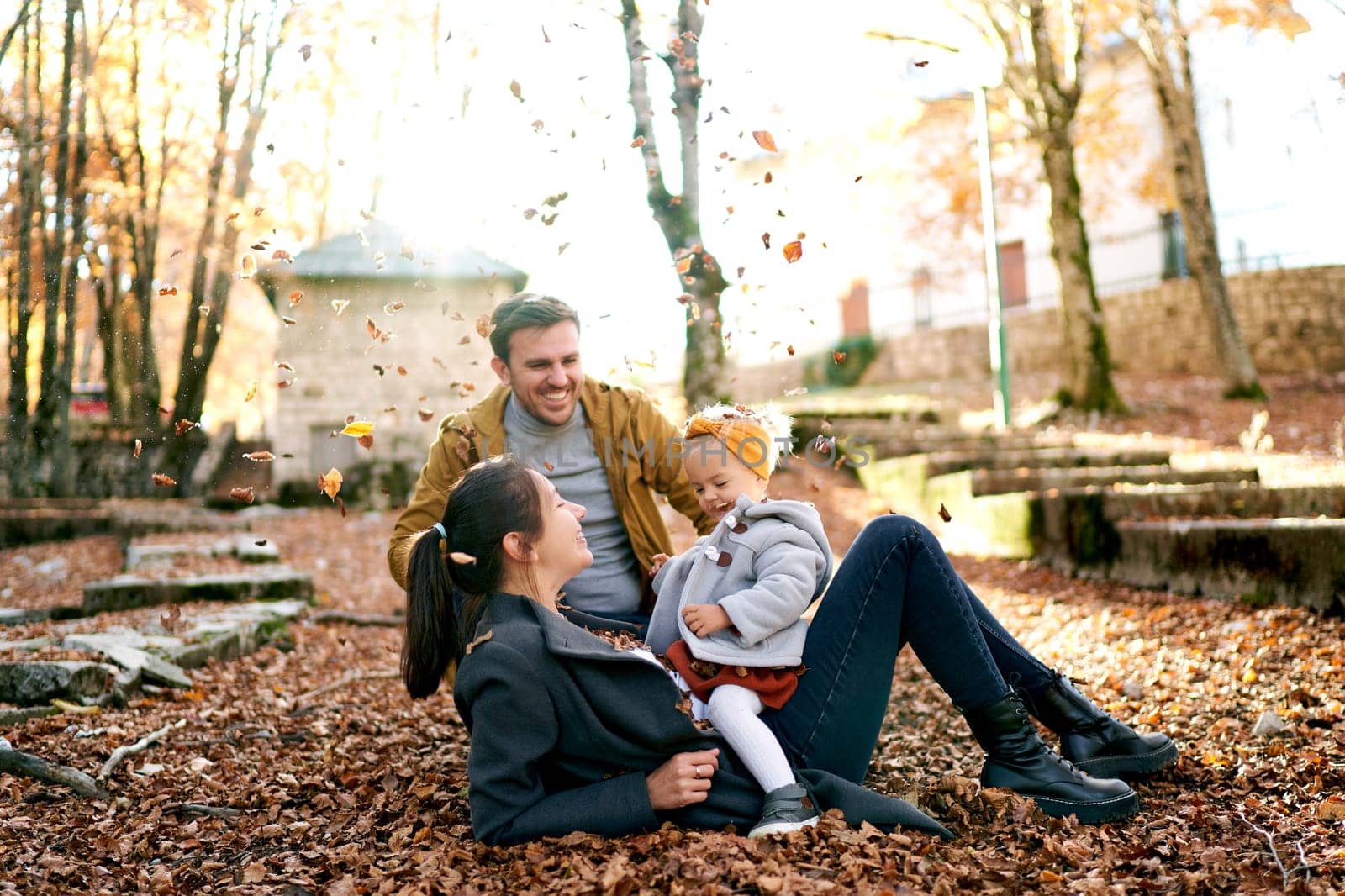 Dad sits next to mom lying on the ground with a little girl on her stomach under falling leaves. High quality photo